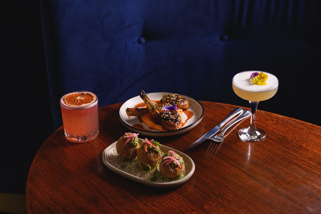 Intimate ONDA table at night — lamb chops on sweet potato puree, golden arancini with green pesto and pickled onions, a grapefruit cocktail with a dried orange wheel, and a foamy egg-white cocktail crowned with edible flowers.