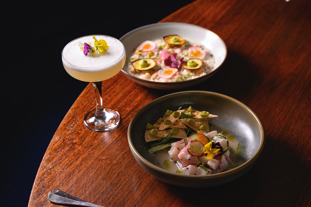 Two ceviche dishes on a dark wooden table — kingfish with pickled radish, edible flowers and bright fish roe; a second white-fish ceviche served with crisp tortilla chips, avocado dollops and a foamy cocktail garnished with violet and yellow blooms.